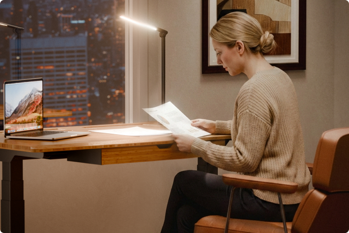Home office scene, woman reading papers at E7 Hive standing desk with desk lamp