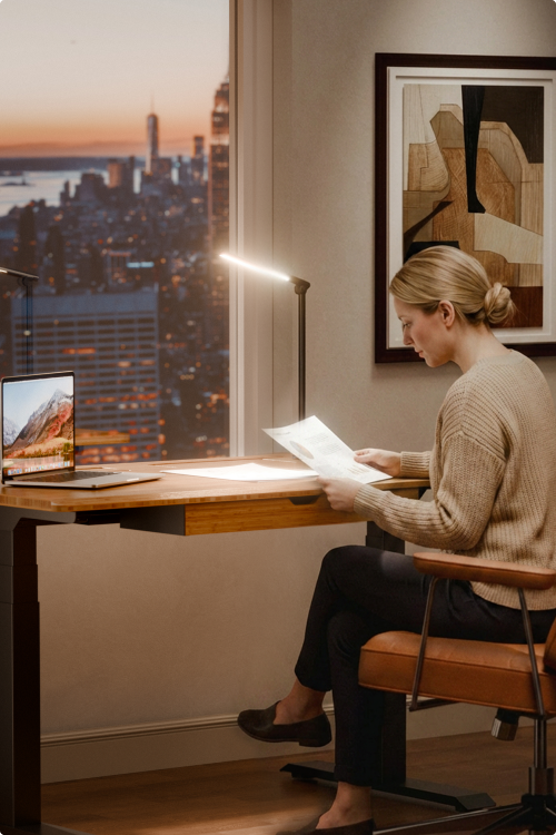 Home office scene, woman reading papers at E7 Hive standing desk with desk lamp