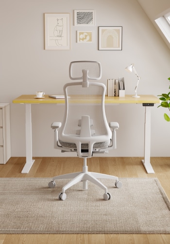 A modern workspace featuring a white ergonomic chair, a yellow desk, a lamp, and framed art on the wall.