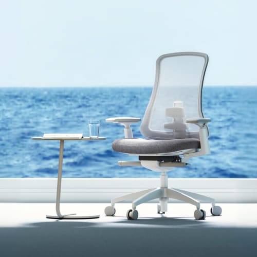 A modern office chair with a mesh back sits beside a small table, overlooking a serene ocean view through a large window.